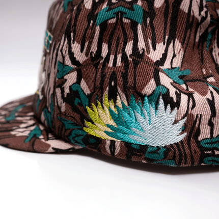 A close-up of a snapback cap featuring a brown, black, and beige tiger-stripe pattern with embroidered teal, light blue, and yellow agave plants on the side panel.